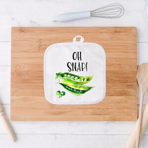 May include: White pot holder with a green pea pod design and the text "OH SNAP!" printed in black.