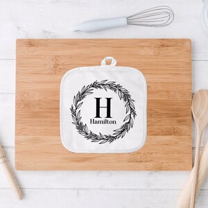 May include: White cotton pot holder with a black floral wreath design and the letter "H" in the center. The text "Hamilton" is below the letter.