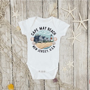 May include: White baby onesie with a graphic of a lighthouse and houses on a beach with the text "Cape May Beach New Jersey, U.S.A."
