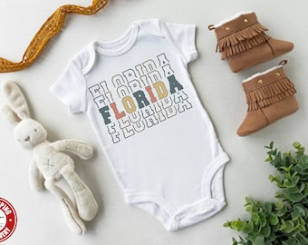 May include: White baby onesie with the word "FLORIDA" repeated in a stacked, retro design in black, orange, yellow, and blue. The onesie is surrounded by a stuffed bunny, a pair of brown suede baby booties, and a green plant.