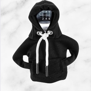 May include: A black shift knob with a white gear pattern, encased in a miniature black hoodie with a white drawstring. The hoodie has a front pocket and metal eyelets.