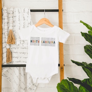 May include: White baby onesie with the text "NORTH CAROLINA" repeated in a stacked, colorful design.