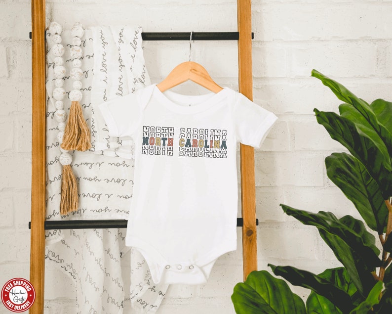 May include: White baby onesie with the text "NORTH CAROLINA" repeated in a stacked, colorful design.