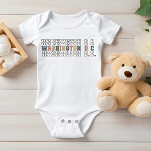 May include: White baby onesie with the text "WASHINGTON D.C." repeated three times in different colors. The text is in a retro font style.