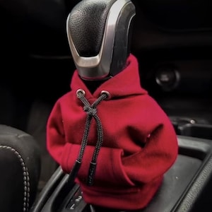May include: A red hooded sweatshirt gear shifter cover for a car. The cover is made of soft fabric and has a drawstring hood.