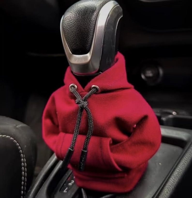 May include: A red hooded sweatshirt gear shifter cover for a car. The cover is made of soft fabric and has a drawstring hood.