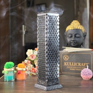 May include: A silver incense tower with a geometric design, emitting smoke. The image also features a Buddha statue, incense sticks, candles, and a box with the text "KULLICRAFT". The items are on a wooden surface.
