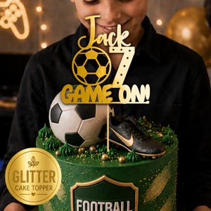 May include: A gold glitter cake topper featuring a soccer ball graphic, the name Jack, the number 7, and the text GAME ON! The topper is mounted on a thin stick and is positioned above a green cake decorated with a soccer ball and a miniature soccer shoe.