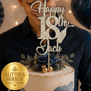 May include: A gold glitter cake topper featuring the text Happy 18th Jack in a cursive and block font combination. The topper is mounted on a thin stick and is displayed on a cake decorated with a dark blue rose and greenery accents.
