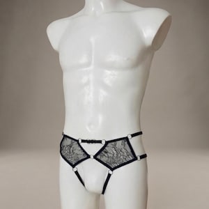 May include: Black lace underwear with a geometric design, displayed on a white mannequin. The underwear features a delicate lace pattern and is accented with black straps and small silver-colored rings.
