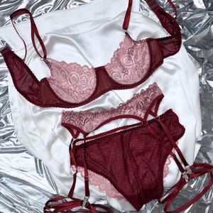 May include: A burgundy lingerie set featuring a bra, panties, and garter belt. The bra and panties have a delicate lace design with a light pink underlay. The set is displayed on a white satin fabric against a silver background.