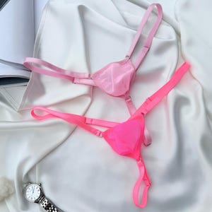 May include: Two sheer thongs in shades of pink, one light and one vibrant, with delicate straps and adjustable details. The lingerie is displayed on a white satin surface, suggesting a focus on intimate apparel.