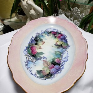 May include: An oval porcelain serving platter with a pink rim and gold trim. The center features a hand-painted design of a floral arrangement with blackberries and leaves.