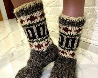 May include: A pair of handmade, thick wool socks. The socks are primarily dark gray with a patterned design in black, white, and burgundy. The cuffs are a lighter color with a textured, fuzzy appearance. The socks appear warm and cozy.