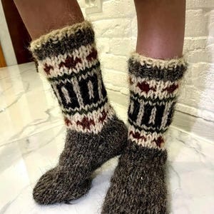 May include: Hand-knitted wool socks in dark grey with a patterned design in black, white, and burgundy. The cuffs are a lighter colour with a textured, fuzzy appearance. These thick socks are designed for warmth and comfort, perfect for cold weather.