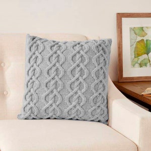May include: A gray knitted throw pillow with a cable knit pattern. The pillow is sitting on a white couch.