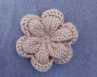May include: A close-up of a knitted flower in a soft pink hue. The flower has six petals and a textured appearance, likely made from yarn. The background is a solid, light purple color.