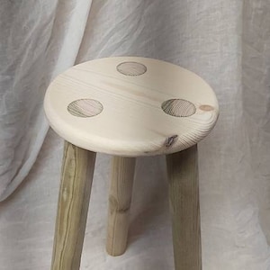 K&ouml;nnte beinhalten: Ein Holzhocker mit drei Beinen und einer runden Oberseite. Die Oberseite hat drei dekorative Kreise.