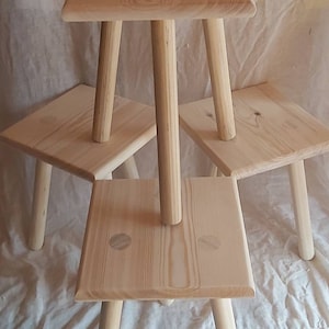 Puede incluir: Cuatro taburetes de madera sin terminar, cada uno con tres patas. Los taburetes están apilados uno encima del otro.