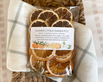 May include: A clear bag containing dried orange slices, chamomile, lemon, and rosemary, labelled "Summer Citrus Simmer Pot." The bag sits in a wicker basket with a cream-coloured cloth. The label has instructions and illustrations of citrus fruits and flowers.