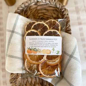 May include: A clear plastic bag filled with dried orange slices, chamomile, lemon, and rosemary, labeled "Summer Citrus Simmer Pot." The bag is nestled in a woven basket with a cream-colored cloth. The label includes instructions and illustrations of citrus fruits and flowers.