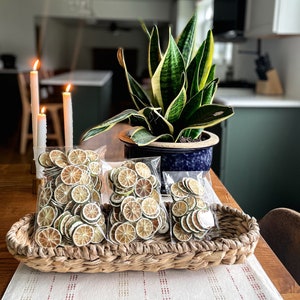 May include: Three bags of dried lime slices in a wicker basket on a wooden table. The lime slices are a light brown color with green edges. The basket is brown and has a woven pattern. There is a green plant in a blue pot in the background.