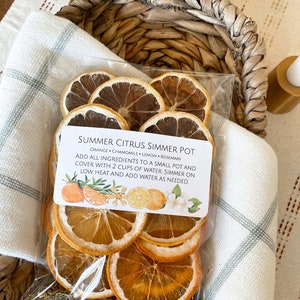 May include: A clear plastic bag filled with dried orange and lemon slices, chamomile, and rosemary, labeled "Summer Citrus Simmer Pot." The bag is nestled in a woven basket atop a white and gray patterned cloth.