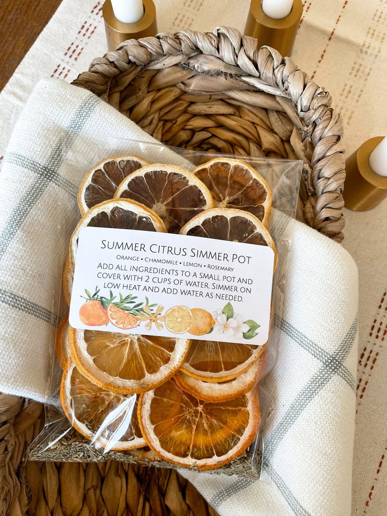May include: A clear plastic bag filled with dried orange and lemon slices, chamomile, and rosemary, labeled "Summer Citrus Simmer Pot." The bag is nestled in a woven basket atop a white and gray patterned cloth.