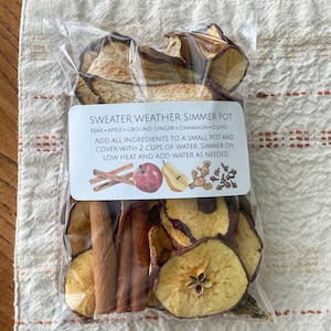 May include: A clear plastic bag filled with dried fruit and spices for a simmer pot. The label reads "Sweater Weather Simmer Pot" and lists ingredients like pear, apple, cinnamon, and cloves. The bag is on a cream-colored cloth.