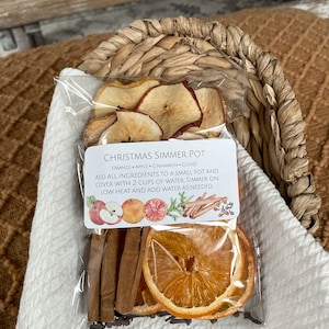 May include: A clear plastic bag containing dried orange slices, apple slices, cinnamon sticks, and cloves. The bag has a label that reads "Christmas Simmer Pot" with instructions. The ingredients are arranged in a woven basket on a white cloth.