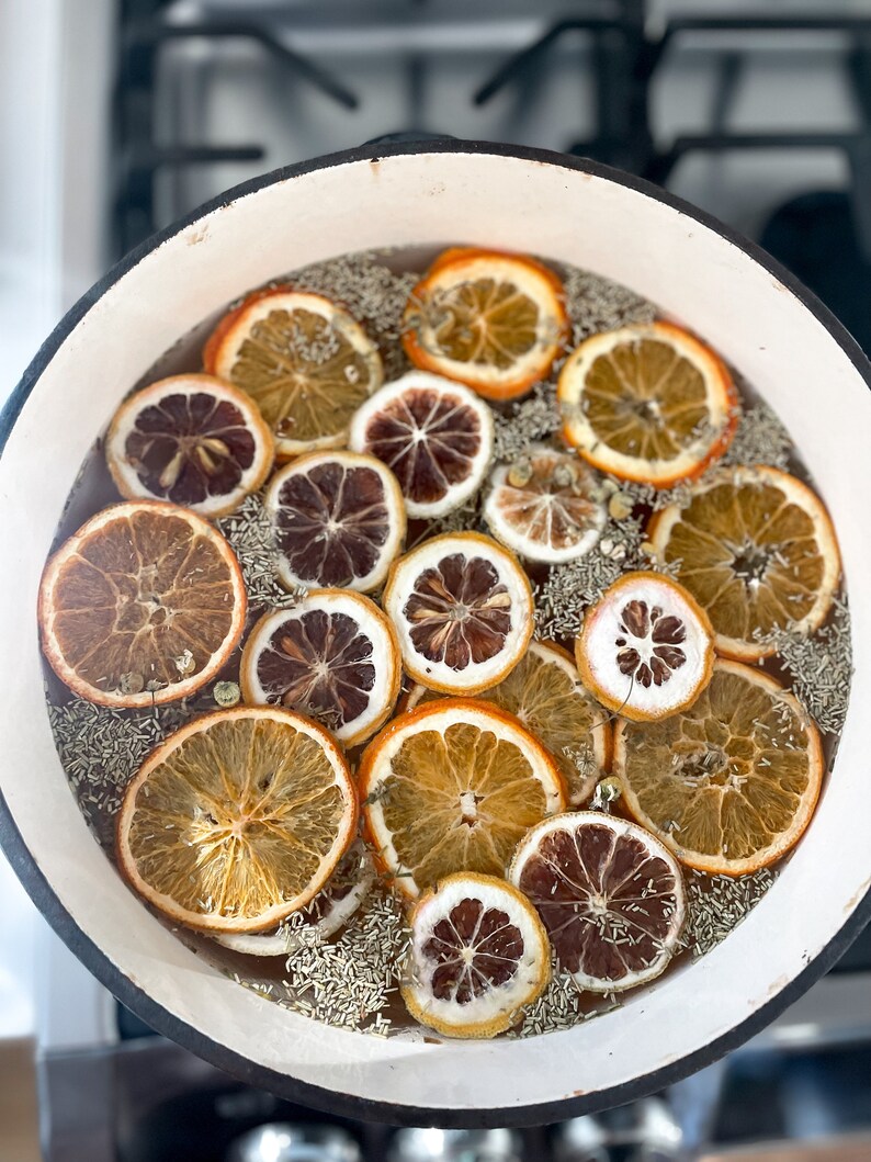 May include: A white enamel pot filled with water, dried orange and lemon slices, and herbs. The orange slices vary in color from light brown to dark brown. The herbs are green and the water is clear. The pot is sitting on a stovetop.