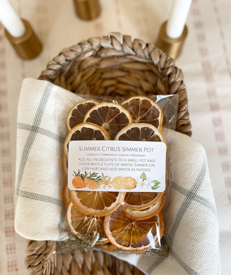 May include: A clear plastic bag filled with dried orange slices, chamomile, lemon, and rosemary, labeled "Summer Citrus Simmer Pot." The bag is nestled in a woven basket with a cream-colored cloth. The label includes instructions and illustrations of citrus fruits and flowers.