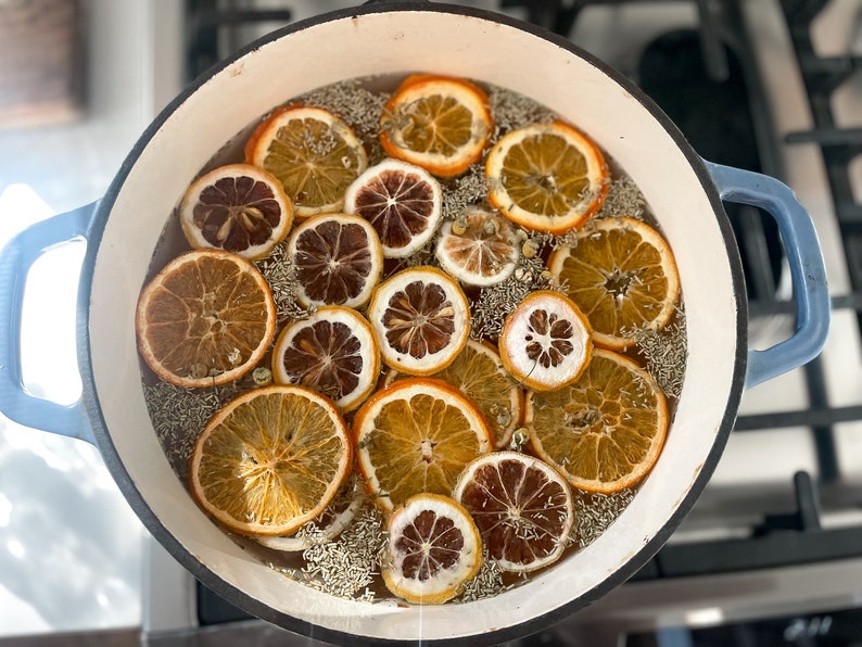 May include: A blue enameled cast iron pot filled with dried orange slices, chamomile flowers, and herbs. The orange slices vary in color from light tan to deep brown. The pot sits on a stovetop.