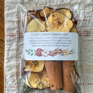 May include: A clear plastic bag containing dried apple slices, cinnamon sticks, thyme, and vanilla bean powder. The label reads "COZY CABIN SIMMER POT" with instructions. The ingredients are for a simmer pot, a home fragrance method.