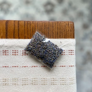 May include: A small, clear plastic bag filled with dried lavender buds. The bag is resting on a cream-colored linen cloth with red and tan stitched stripes. The background includes a wooden surface.