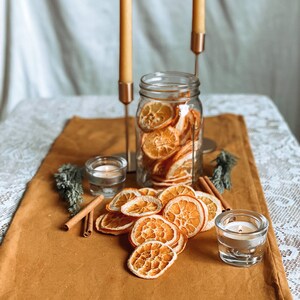 May include: A glass jar filled with dried orange slices sits on a brown table runner. Two clear glass candle holders with lit candles are on the table. The jar and candle holders are surrounded by dried orange slices and cinnamon sticks.