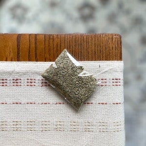 May include: A small clear plastic bag filled with dried rosemary leaves. The bag is sitting on a white and red striped fabric.