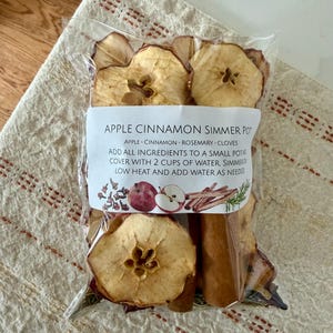 May include: A clear plastic bag containing dried apple slices, cinnamon sticks, and other ingredients for a simmer pot. The label reads "Apple Cinnamon Simmer Pot" with instructions. The bag is on a beige and red patterned cloth.