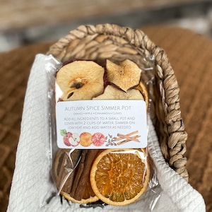 May include: A clear plastic package containing dried orange slices, apple slices, cinnamon sticks, and cloves, labeled "Autumn Spice Simmer Pot." The package is nestled in a woven basket atop a white cloth.