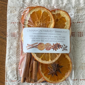 May include: A clear plastic bag containing dried orange slices, cinnamon sticks, star anise, and cloves. The label reads "CINNAMON HARVEST SIMMER POT" with instructions. The ingredients are arranged on a beige cloth.