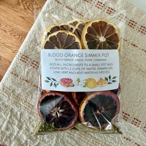 May include: A clear plastic bag filled with dried blood orange slices, thyme, and other ingredients. The label reads "Blood Orange Simmer Pot" with instructions. The bag is on a beige and white patterned cloth.