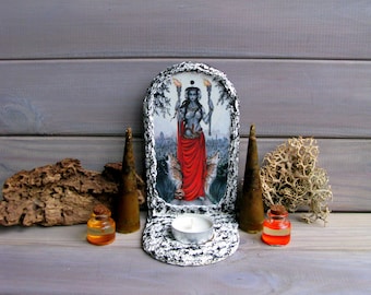 May include: A wooden altar with a white painted finish and a black and white image of a woman holding torches. The altar has a candle holder and two small bottles with orange liquid.