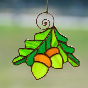 May include: A stained glass ornament featuring a green oak leaf with two acorns. The acorns are yellow and green.