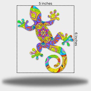 May include: A decorative lizard with a detailed, patterned design. The lizard is approximately 12.7 cm wide and 15.2 cm tall. The design features yellow, green, blue, purple, and orange. A vibrant piece of art.