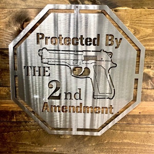 May include: A silver metal sign with an octagonal shape. The sign has the text "Protected By The 2nd Amendment" and a silhouette of a handgun.
