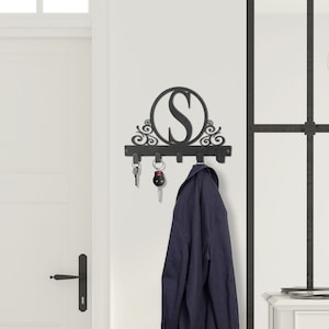 May include: Black metal key rack with five hooks and a decorative circle with the letter "S" in the center. The rack is mounted on a white wall and has a coat hanging on one of the hooks.