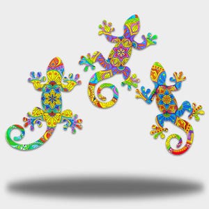 May include: Three decorative gecko ornaments in vibrant colours. Each gecko has a detailed, patterned design in yellow, blue, green, orange, and purple. The lizards have curled tails and are set against a white backdrop.