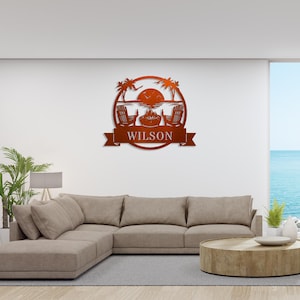 May include: A metal wall art piece with a circular design featuring two beach chairs, a bonfire, palm trees, and a sunset. The text "WILSON" is displayed on a banner below the circle.