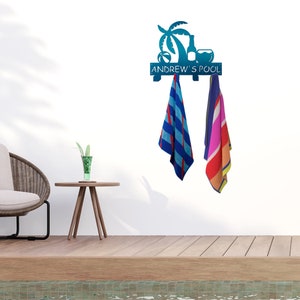 May include: A teal metal pool towel rack with a palm tree, bottle, and beach ball design. The text "ANDREW'S POOL" is displayed on the rack. Two colorful striped towels hang from the rack, with a chair and table in the background.