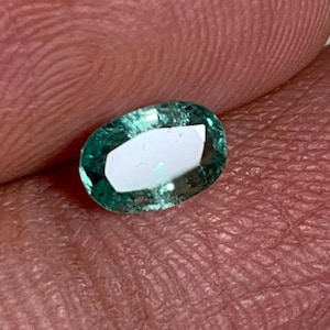 May include: An oval-shaped, teal-colored gemstone with a bright, white interior. The gem is held between two fingers, showcasing its polished surface and clarity. The gemstone's color is a vibrant teal, with a reflective, polished surface.